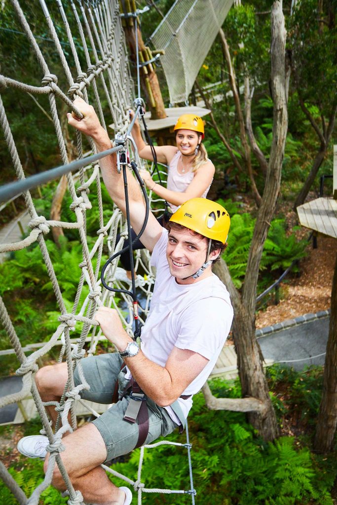 Pair of adults smile at camera while hanging from the net wall obstacle on the Grand Tree Surfing course at Enchanted Adventure