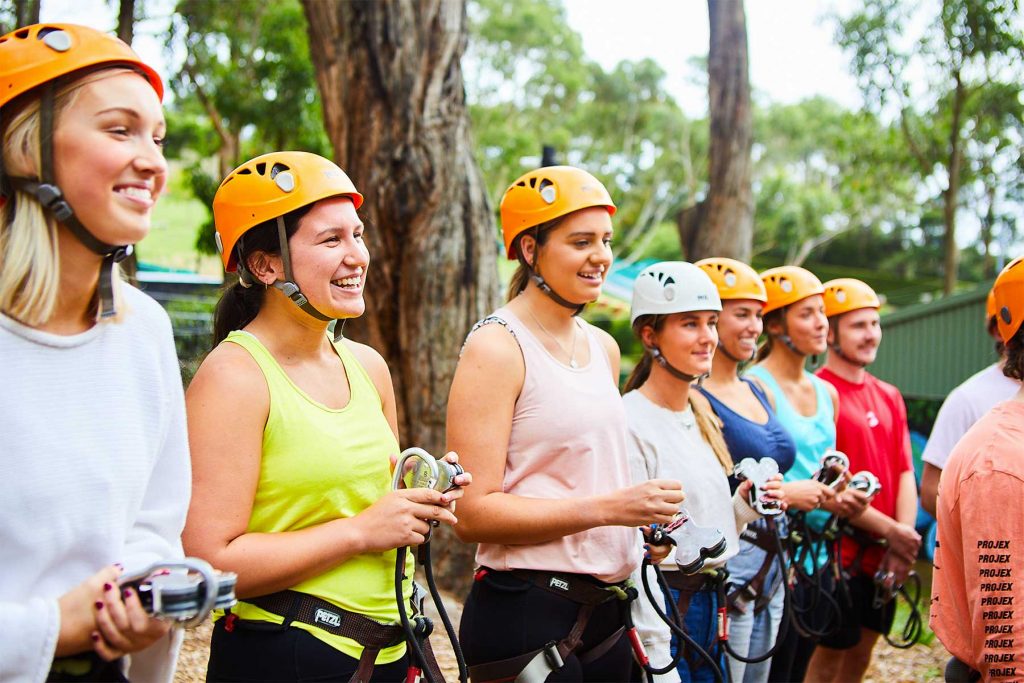 Group of young adults watching our experienced instructor perform the safety brief demonstration for the Grand Tree Surfing course at Enchanted Adventure